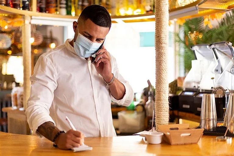Hombre con máscara, tomando pedidos telefónicos y escribiendo en un bar.