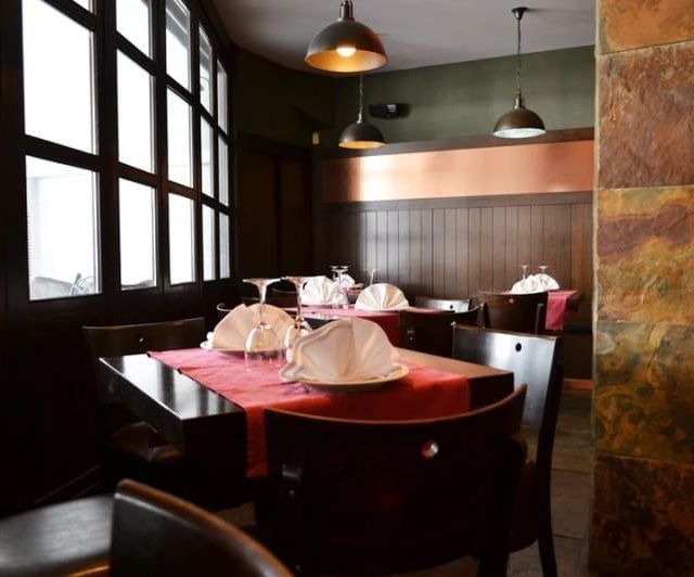Comedor de restaurante con mesas preparadas para el servicio. Muebles de madera oscura, detalles en rojo y pared de piedra.