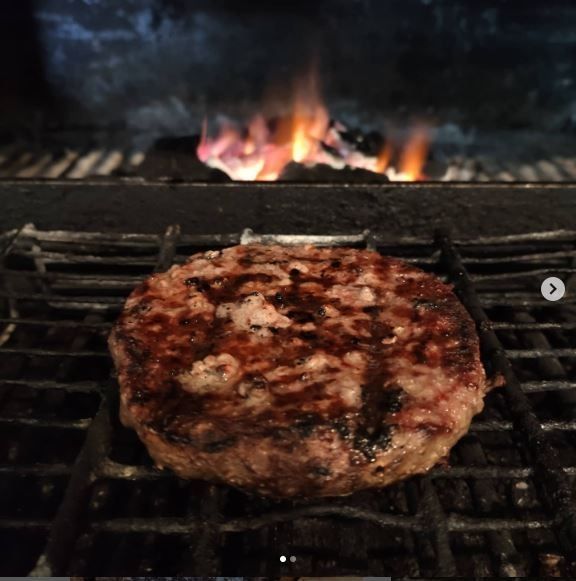 Una hamburguesa asándose en una parrilla oscura alimentada por carbón con llamas visibles en el fondo.