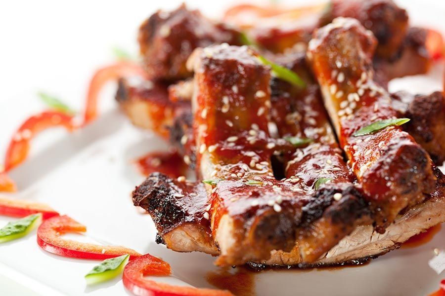 Costillas a la parrilla con semillas de sésamo y salsa, adornadas con pimientos rojos en un plato blanco.