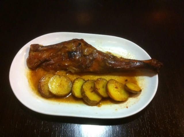 Pierna de cordero asada con patatas en rodajas en un plato ovalado blanco.