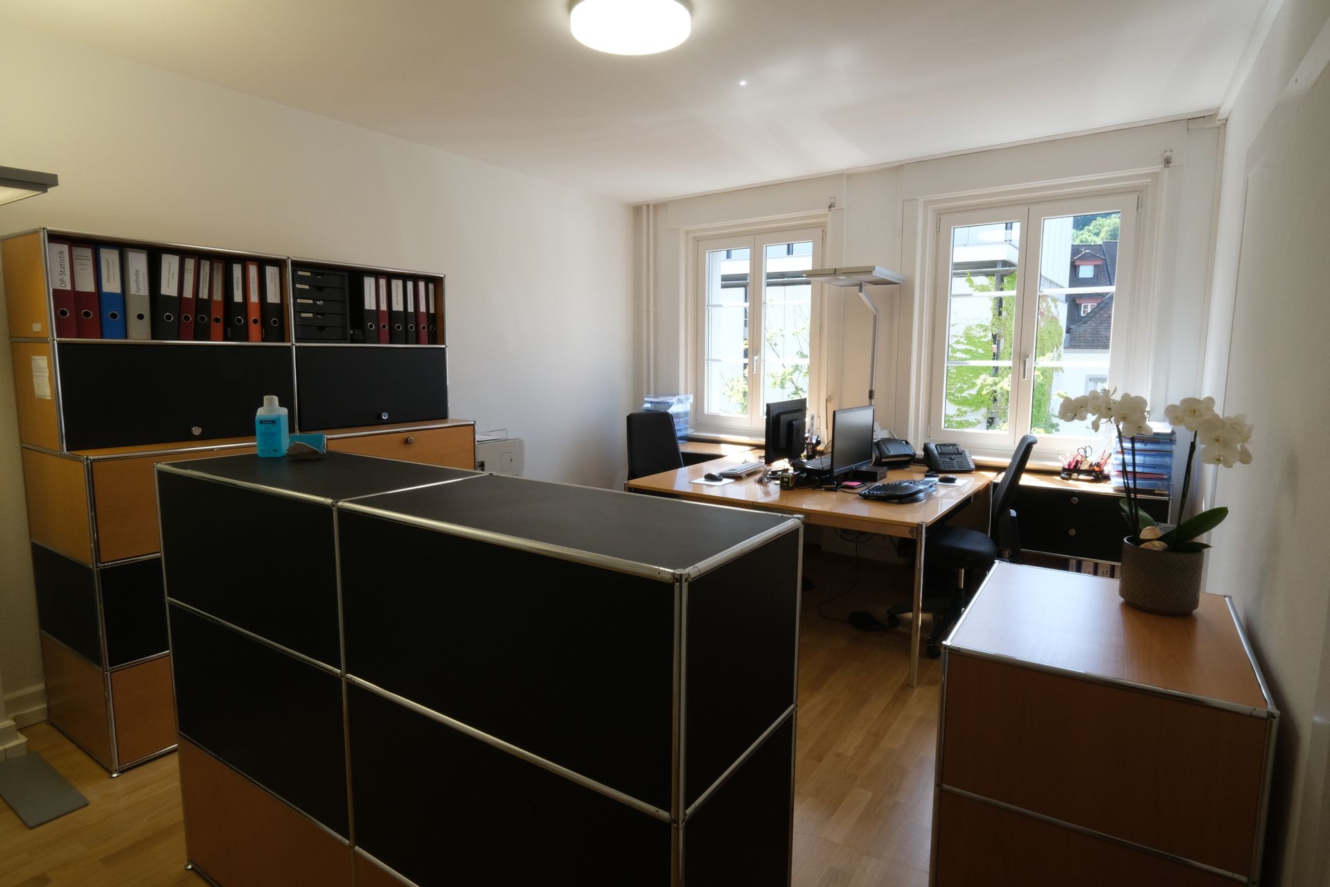 Ein Zimmer mit einem Schreibtisch, Stühlen, Regalen und einer Uhr an der Wand.