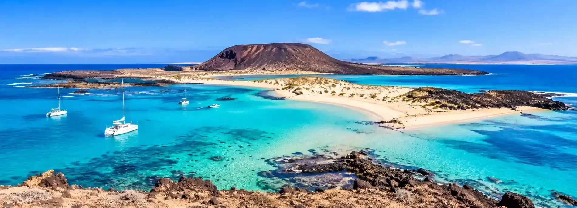 Bahía de color turquesa con veleros, islas arenosas y una colina rocosa bajo un cielo azul brillante