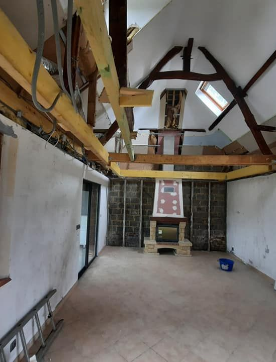 Rénovation intérieure : poutres apparentes, cheminée et murs non finis dans une pièce avec plafond mansardé et puits de lumière.