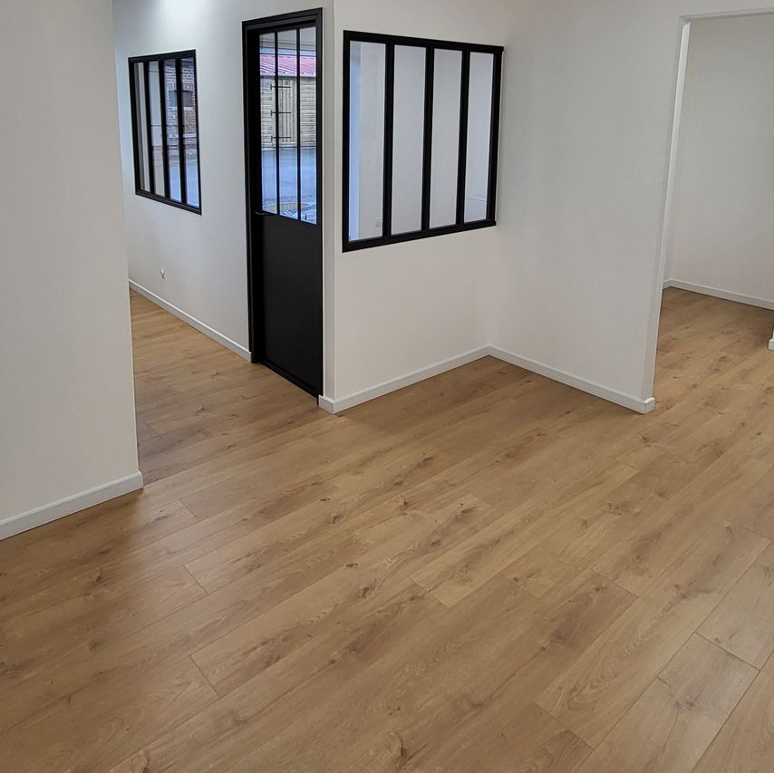 Pièce vide avec parquet, murs blancs, fenêtres et porte à cadre noir.