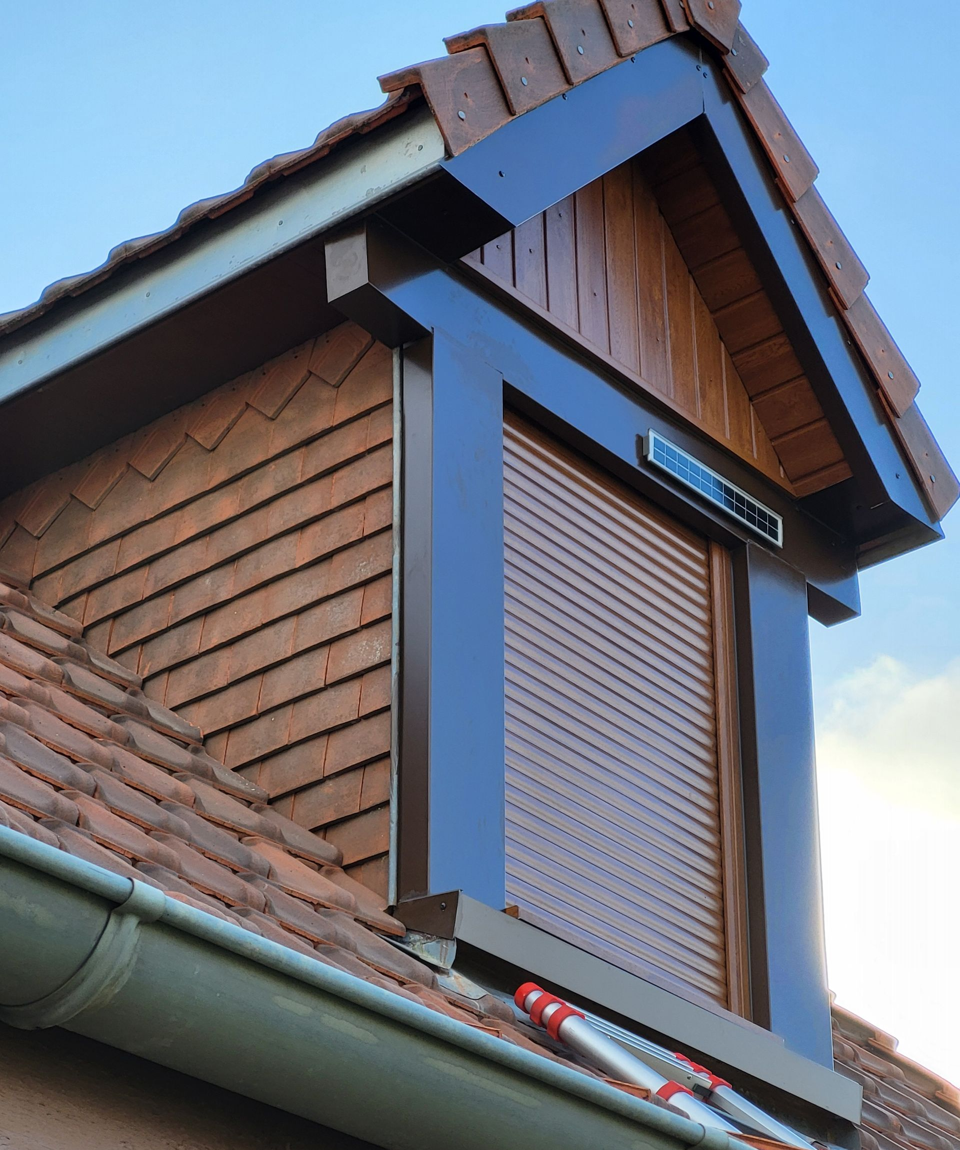 Lucarne brune avec volet roulant fermé sur un toit de tuiles.