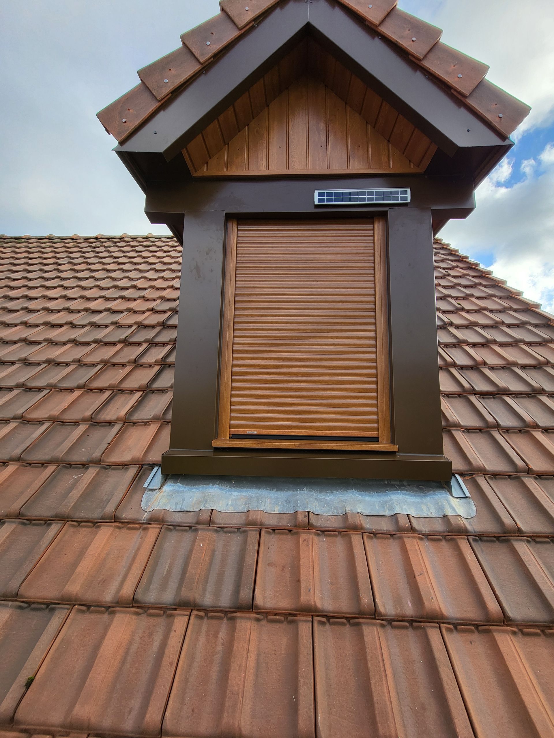 Puits de lumière à cadre brun et volets fermés sur un toit de tuiles.