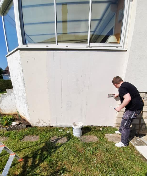 Une personne applique du plâtre sur un mur extérieur jouxtant une structure à toit de verre.