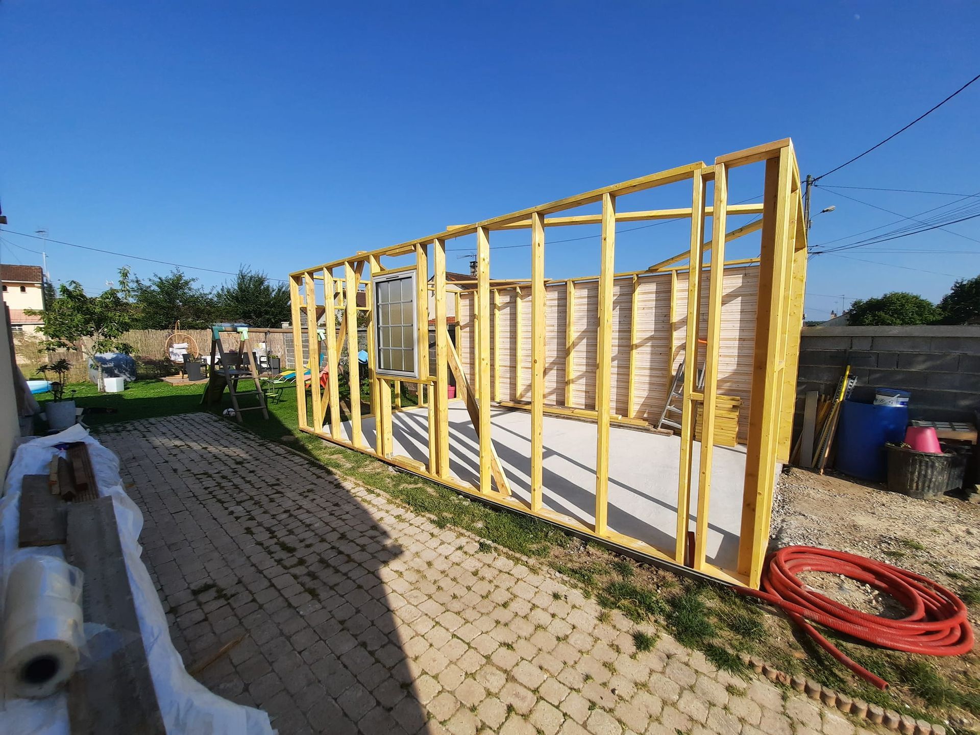 La charpente en bois d'un bâtiment en construction, posée sur une terrasse pavée par une journée ensoleillée.