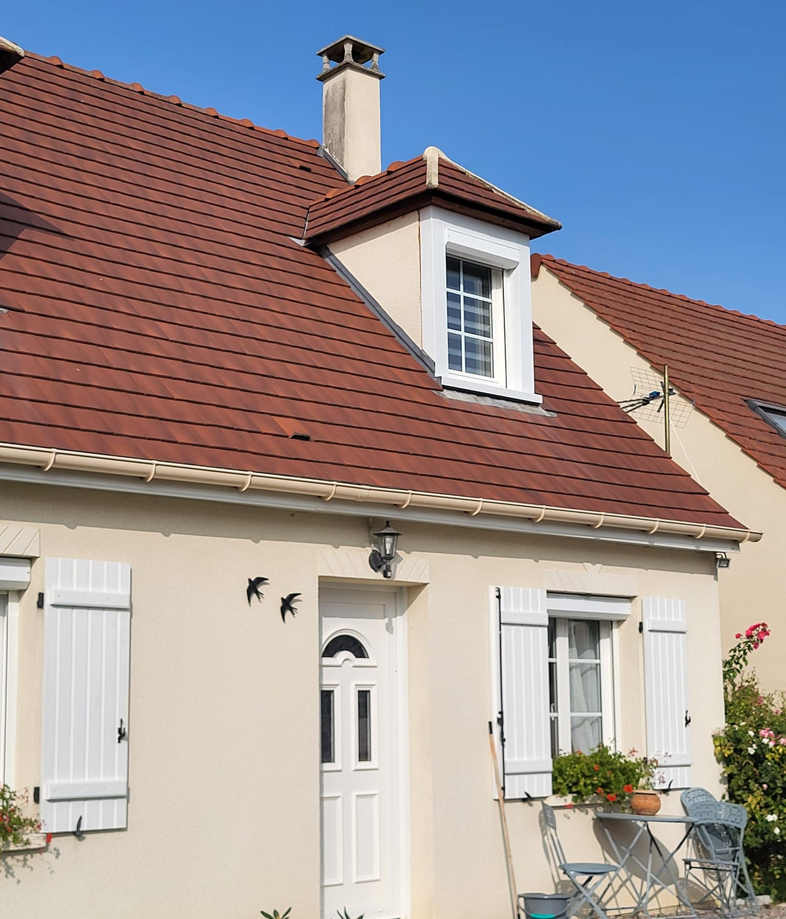 Maison beige au toit rouge, volets blancs et lucarne. Ciel bleu.