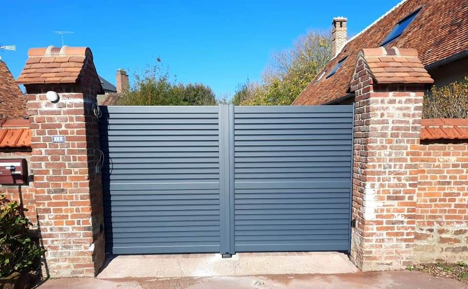 Portail à lattes horizontales grises entre des piliers de briques.