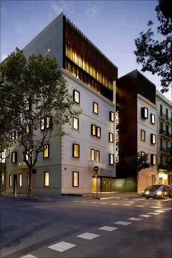 Exterior moderno de un hotel al atardecer, fachada gris, ventanas iluminadas, coche en la calle.