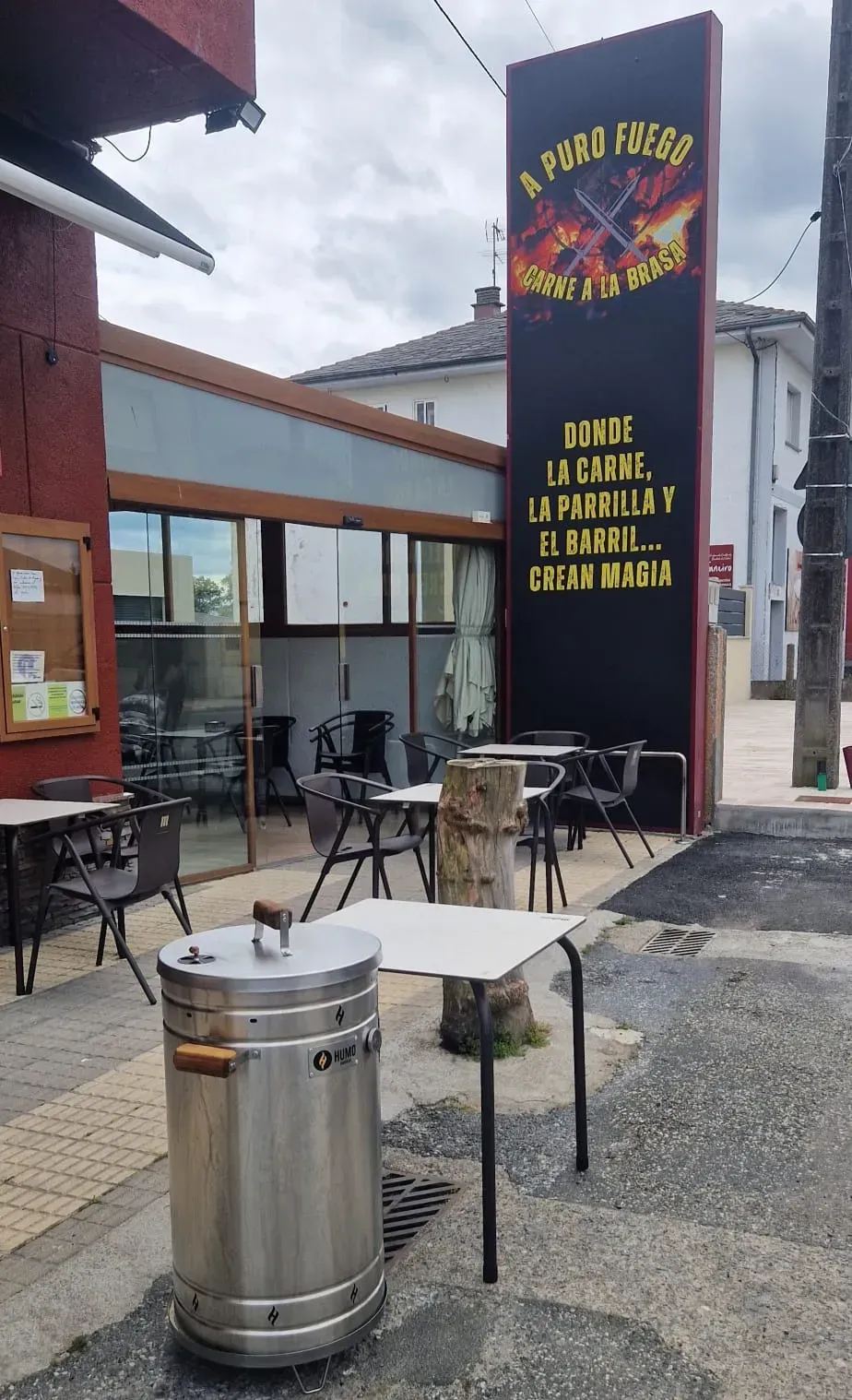 Mesa de café al aire libre junto a un letrero negro de restaurante y un cubo de basura de acero inoxidable en un patio de ladrillos.