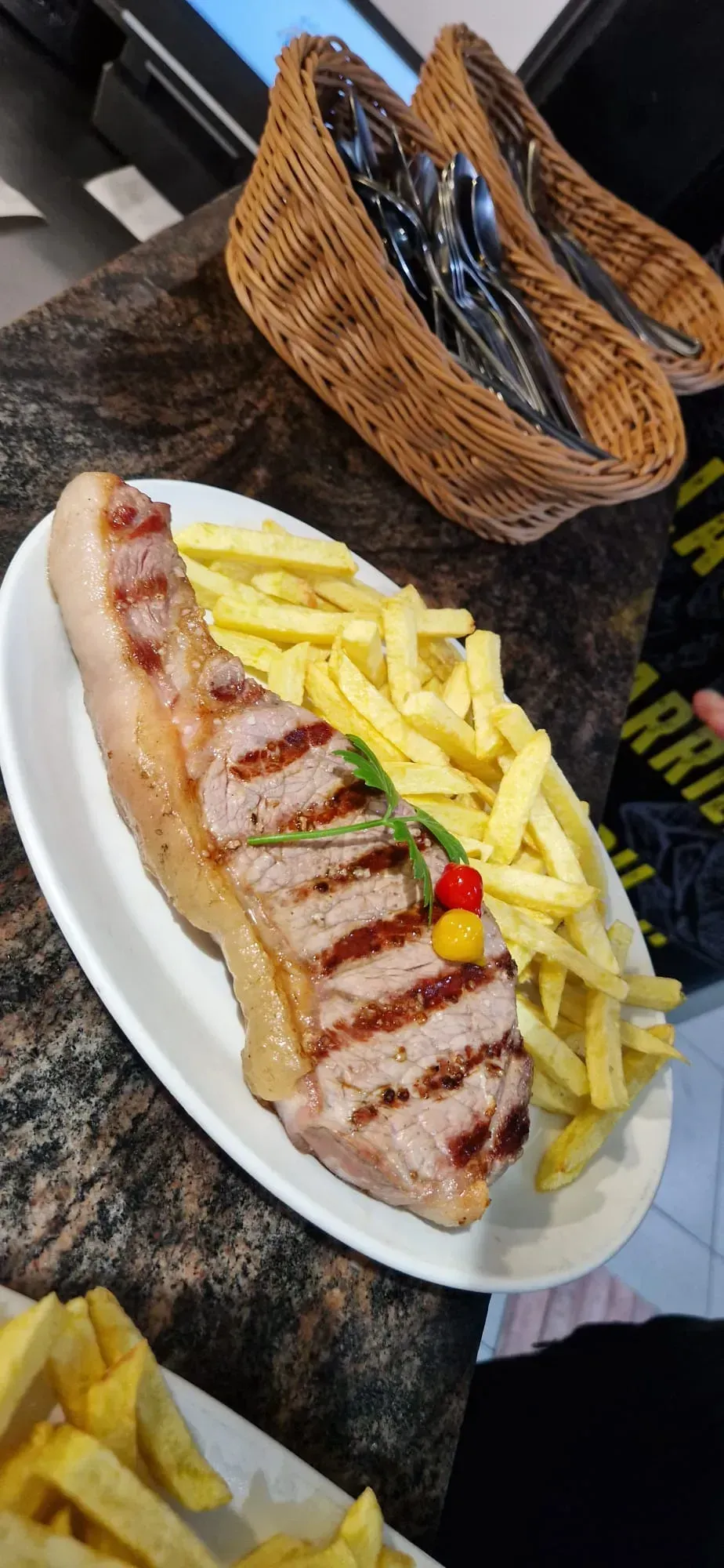 Sándwich de pollo a la parrilla con papas fritas en un plato blanco, con una cesta de mimbre llena de papas fritas al fondo.