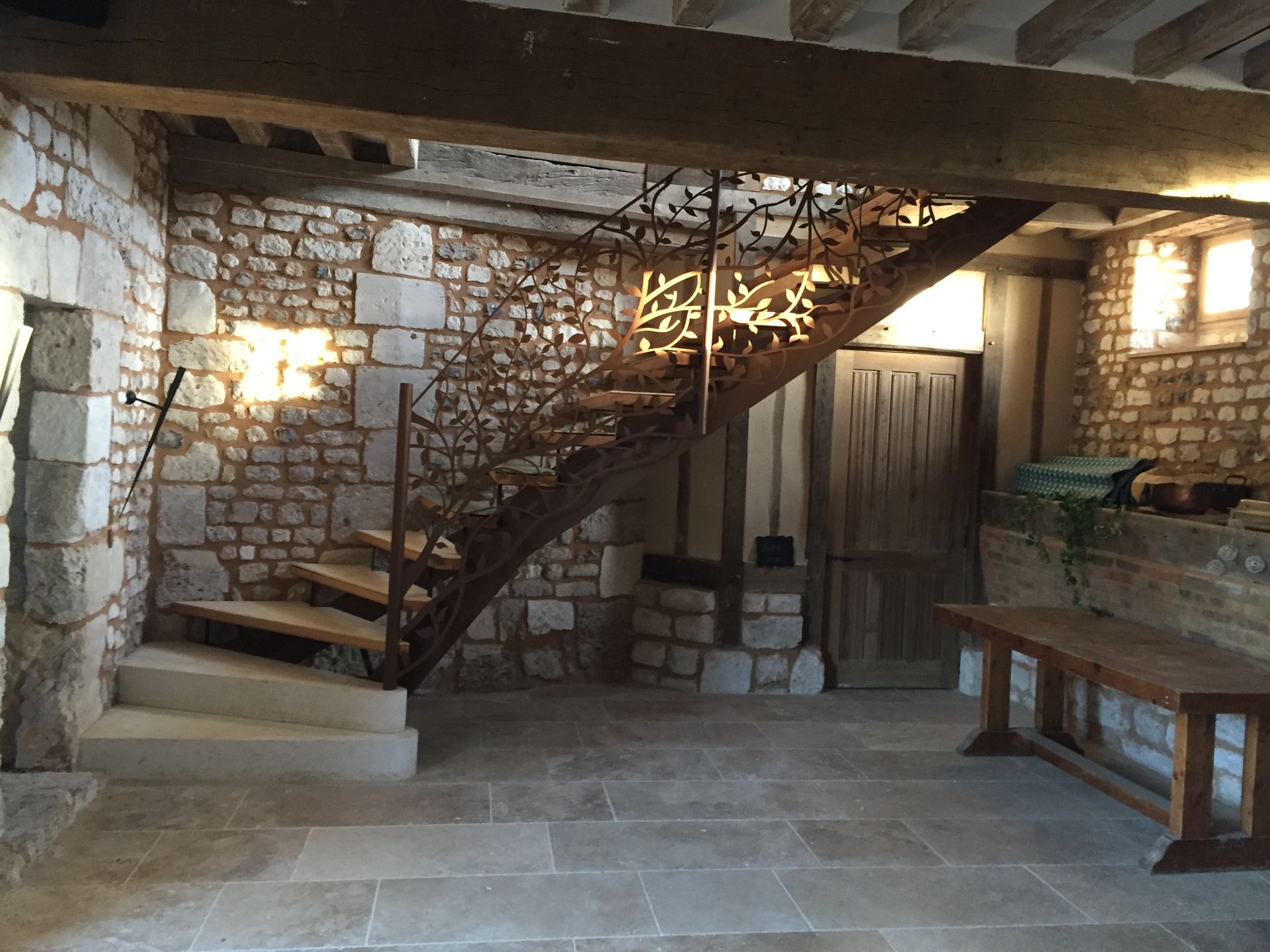 Intérieur en pierre avec escalier en colimaçon, porte en bois et longue table à manger éclairée par des appliques murales chaleureuses.