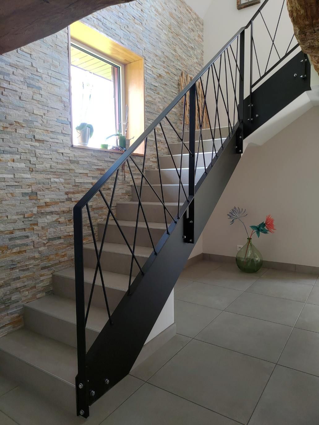 Escalier intérieur avec rampe en métal noir, mur en pierre, sol carrelé et fenêtre laissant entrer la lumière.