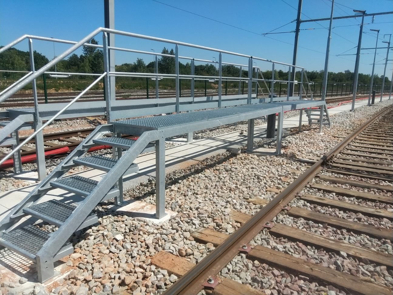 Passerelle piétonne métallique enjambant une voie ferrée sur un chantier de construction sous un ciel bleu dégagé