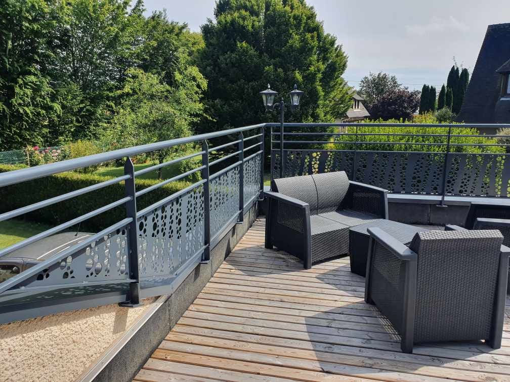 Terrasse extérieure avec sièges en osier gris, parquet et rambarde en métal donnant sur la verdure.