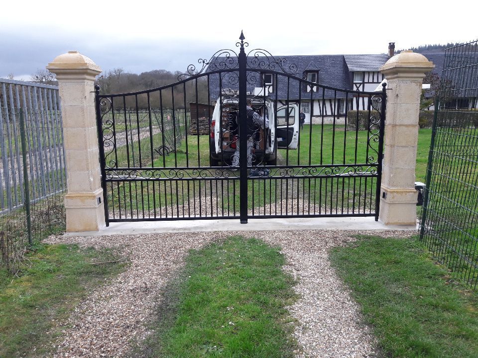 L'entrée du jardin est fermée par un portail en fer noir, des piliers en pierre et un chemin menant à une pelouse et un kiosque à l'intérieur.