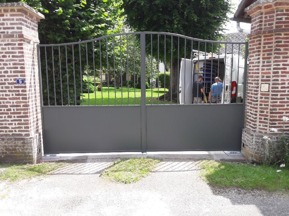 Un portail métallique noir fermé, entre des piliers de briques, laisse entrevoir deux personnes à l'intérieur, sur un chemin de jardin.