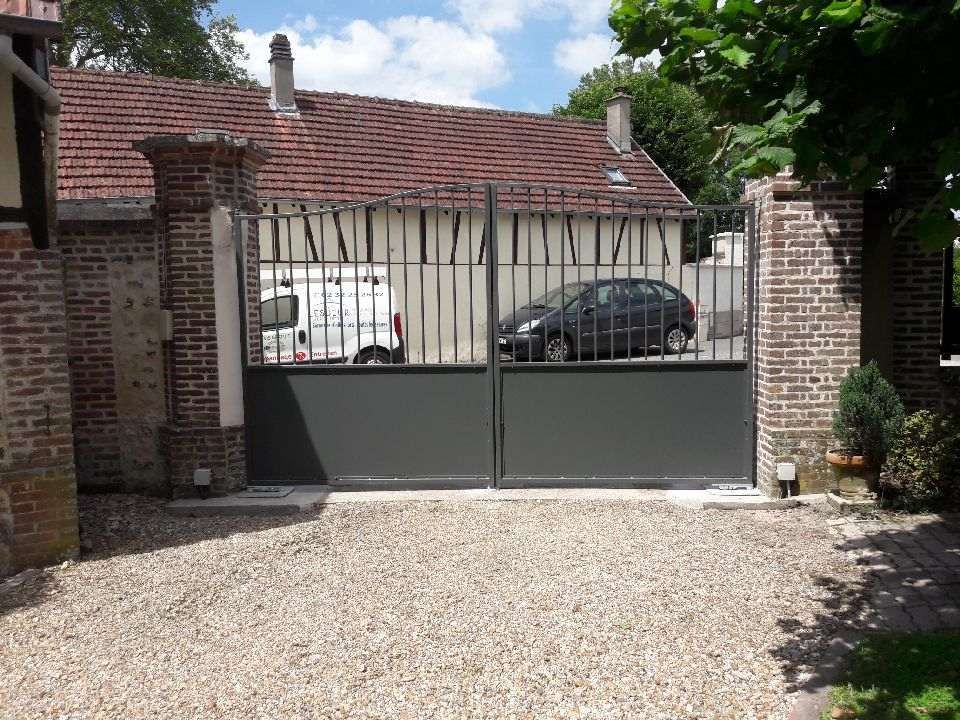 Portail d'allée gris fermé dans un mur de briques, avec des voitures garées et une maison derrière.