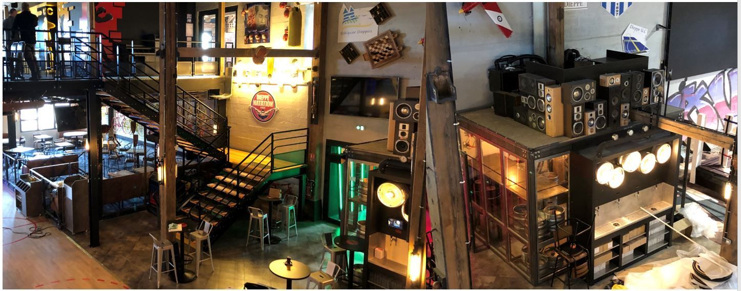 Intérieur de loft industriel sur deux étages avec escalier en métal, éclairage chaleureux et étagères remplies d'appareils électroniques et d'objets de décoration.