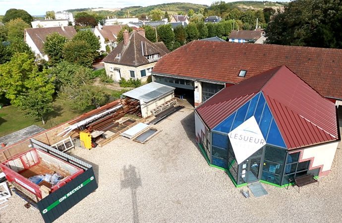 Vue aérienne d'un terrain gravillonné jouxtant un bâtiment au toit rouge et plusieurs maisons, avec des arbres et des conteneurs stationnés.