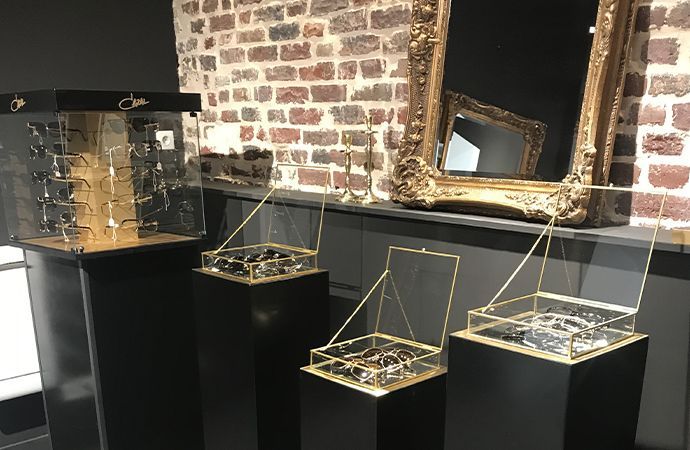 Vitrines géométriques en verre sur socles noirs dans une galerie aux murs de briques avec un grand miroir orné.