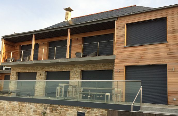 Maison moderne à deux étages avec bardage en bois, soubassement en pierre et balustrade de balcon en verre.