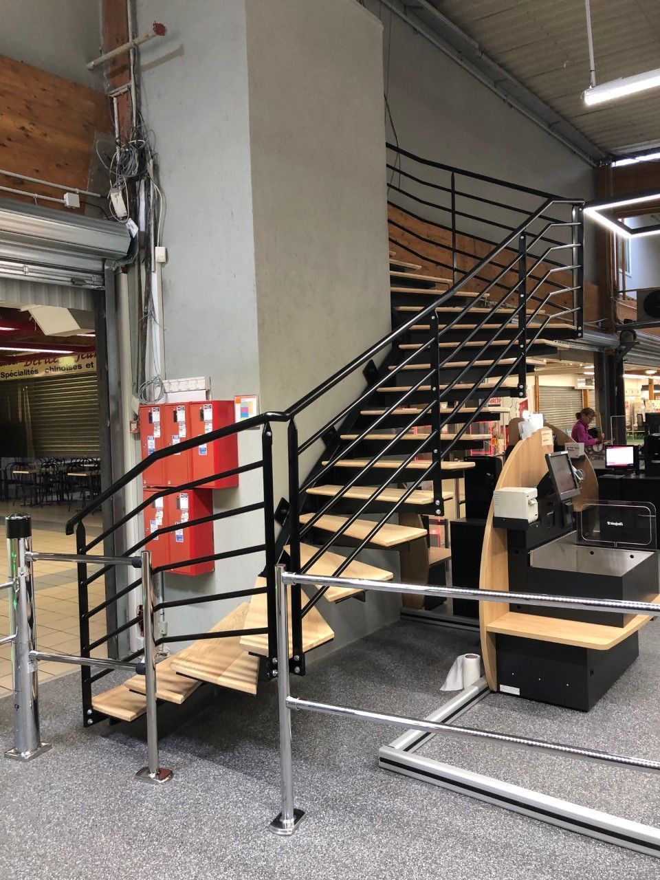 Bureau aménagé dans un entrepôt, avec un escalier noir, une rampe en métal, un extincteur rouge et des bureaux sous l'escalier.