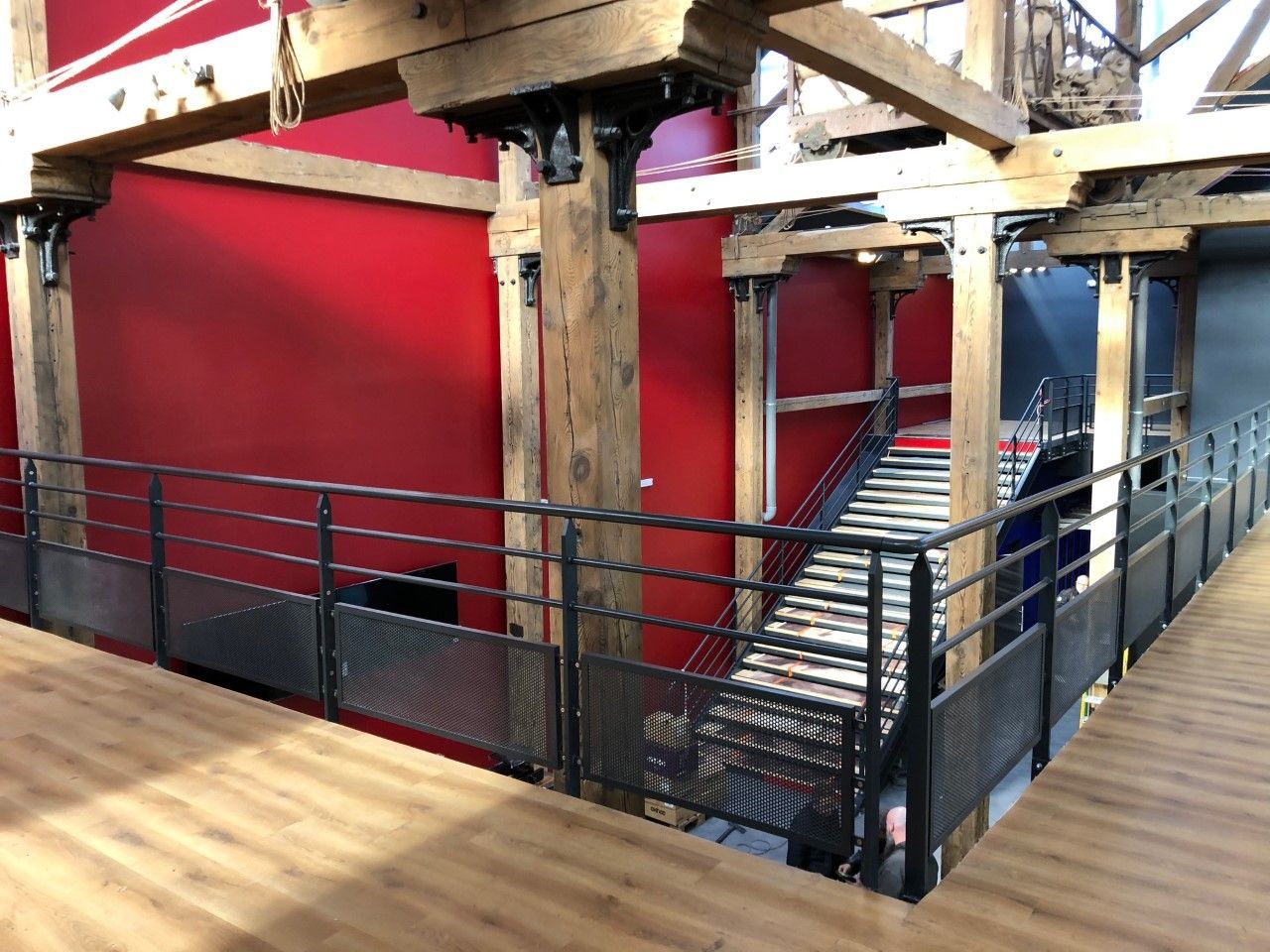 Balcon intérieur avec balustrades noires, poutres en bois et murs rouges donnant sur un escalier situé au niveau inférieur.