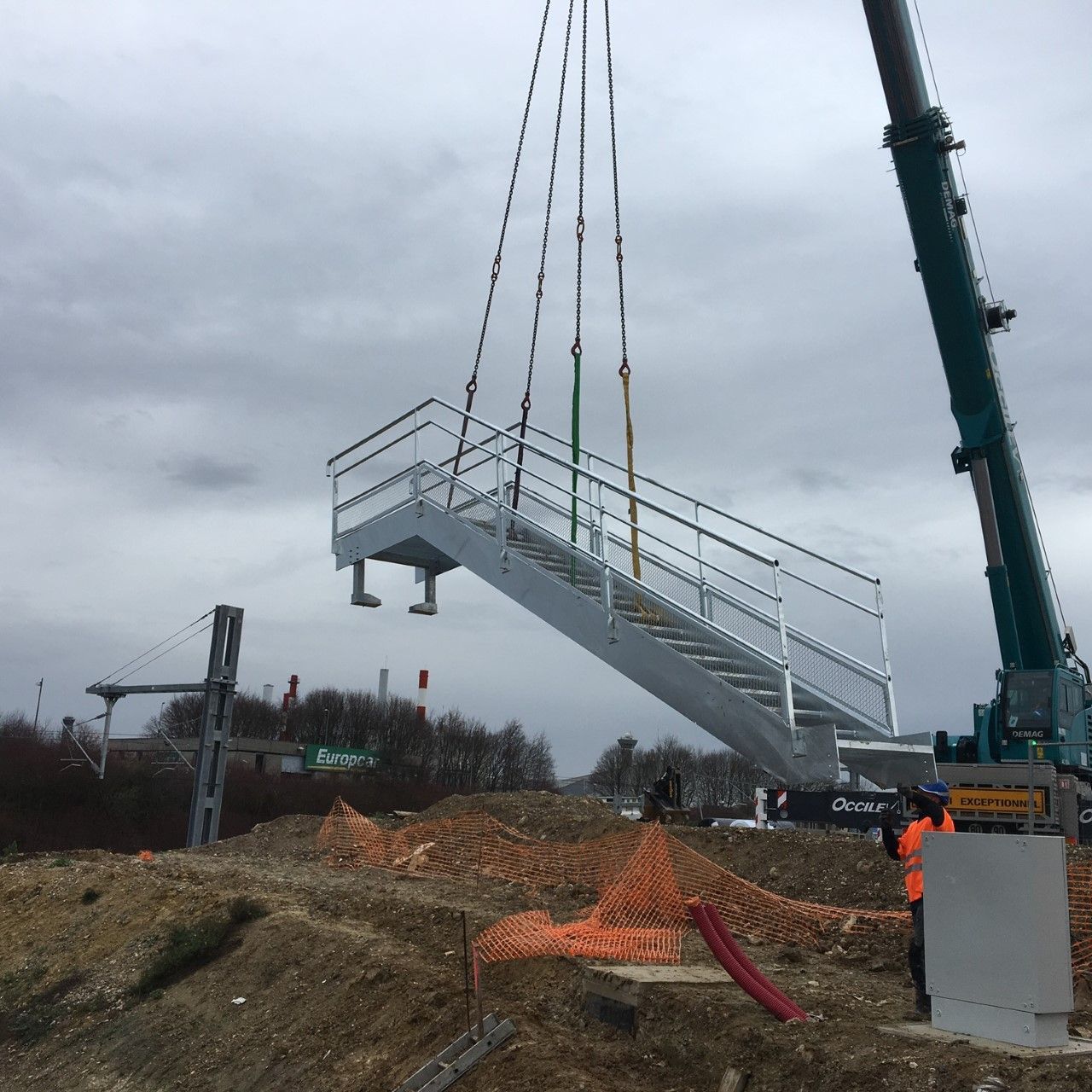 Une grue soulève un escalier métallique blanc sur un chantier boueux sous un ciel gris.
