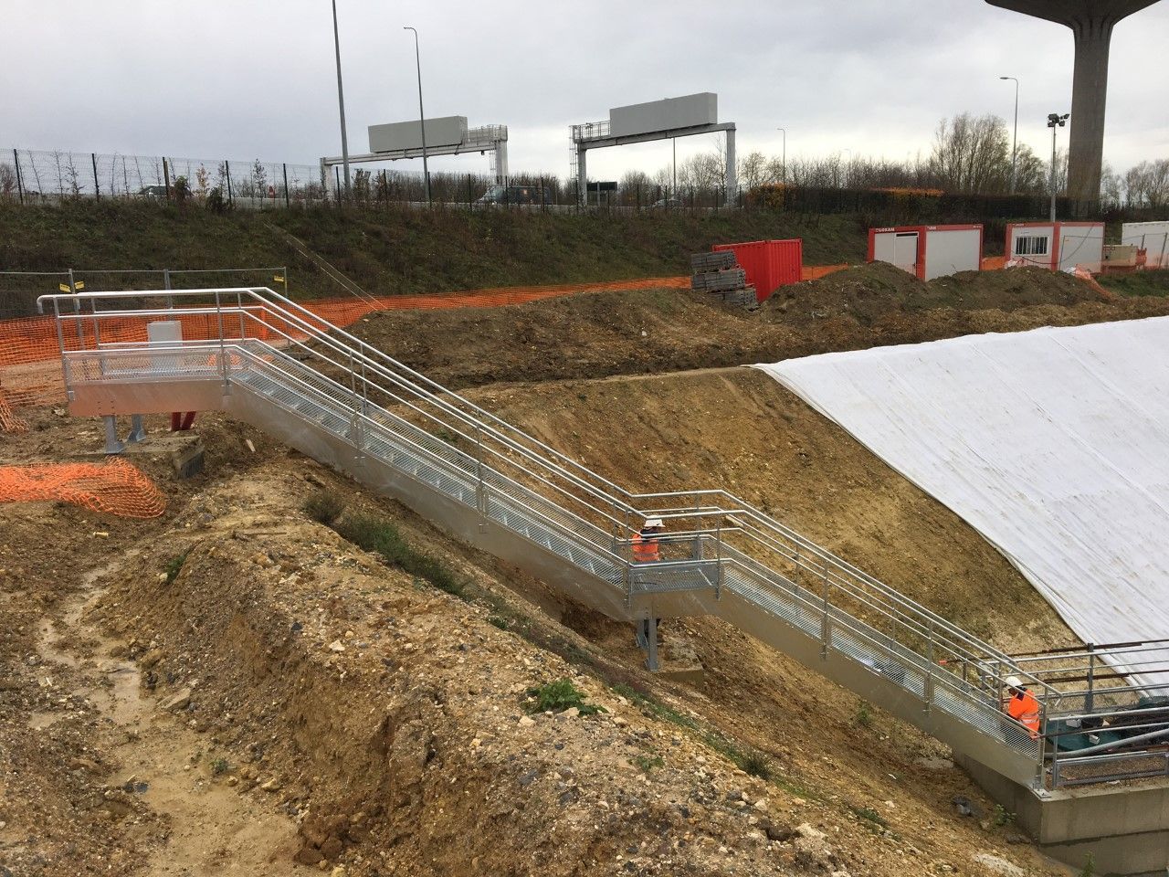 Chantier de construction extérieur avec un canal en béton et des garde-corps métalliques à côté d'un talus de terre en pente.