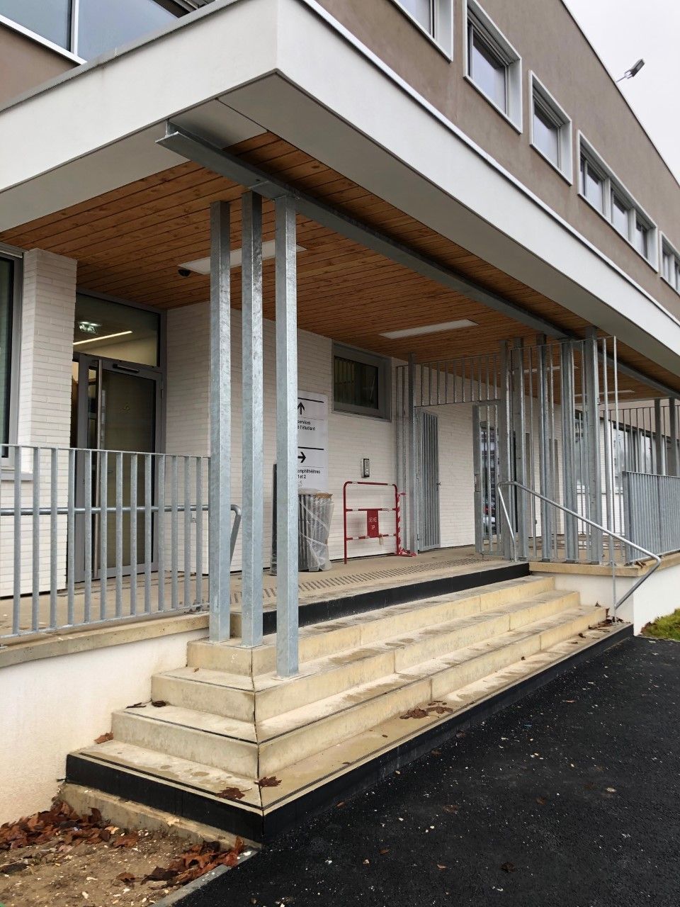 Entrée du bâtiment avec marches en béton, rambardes métalliques et entrée couverte à côté d'un chemin asphalté mouillé
