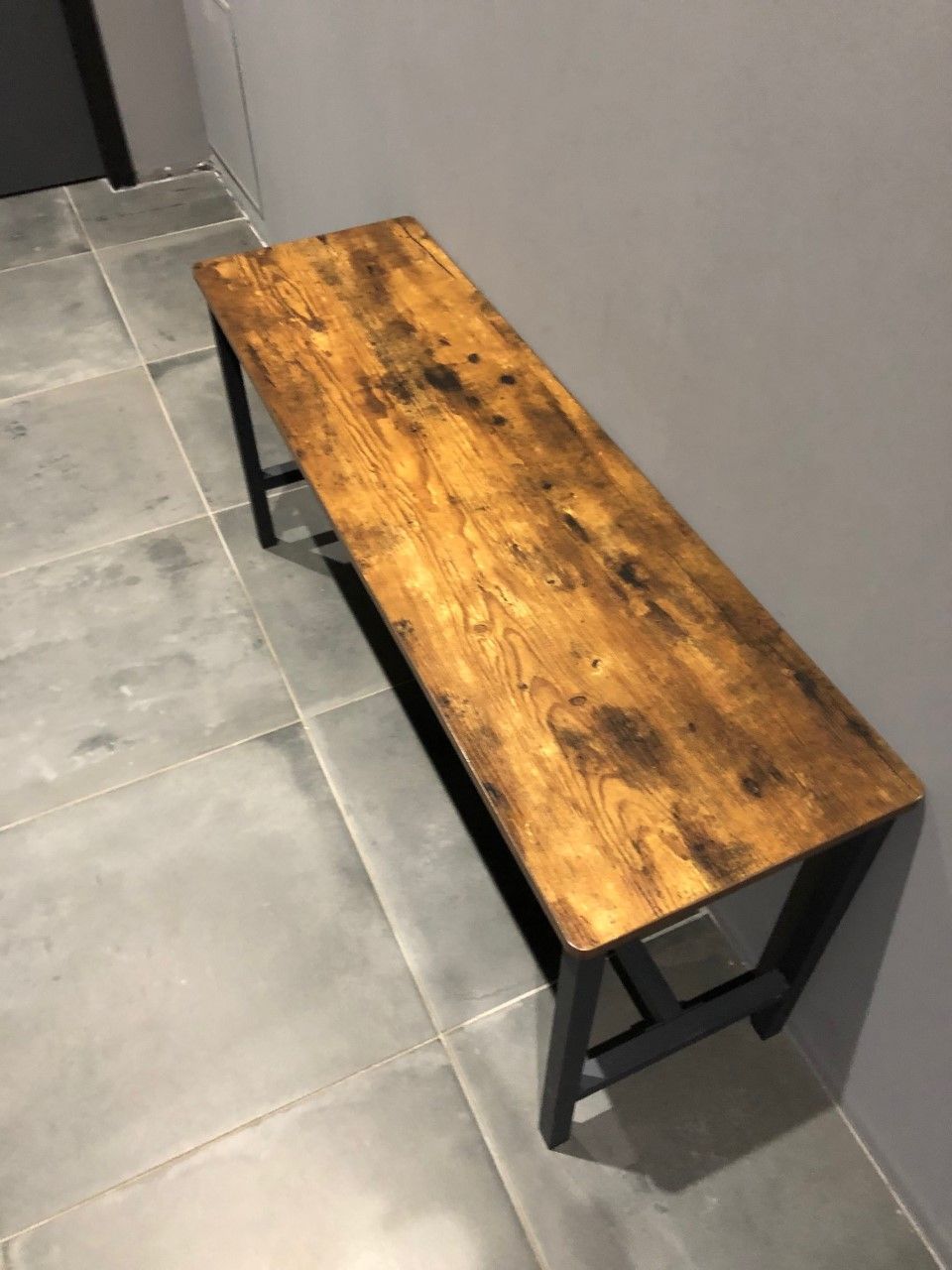 Banc rustique en bois avec pieds en métal noir contre un mur gris sur un sol carrelé