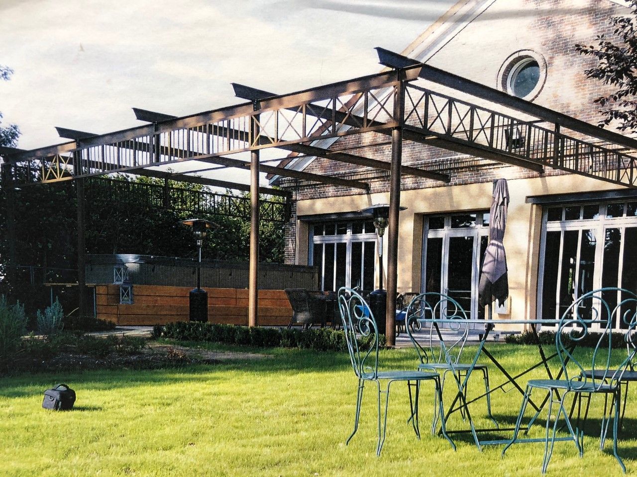 Terrasse de café en plein air avec tables et chaises en métal sous une pergola à côté d'un bâtiment, avec pelouse verte et arbres.