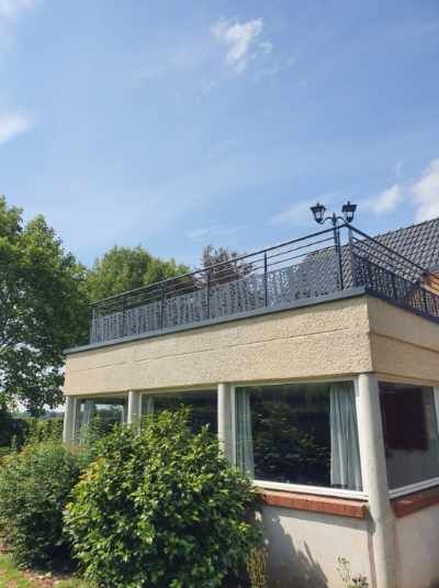 Petit bâtiment beige avec de grandes fenêtres et une terrasse sur le toit sous un ciel bleu, entouré d'arbres
