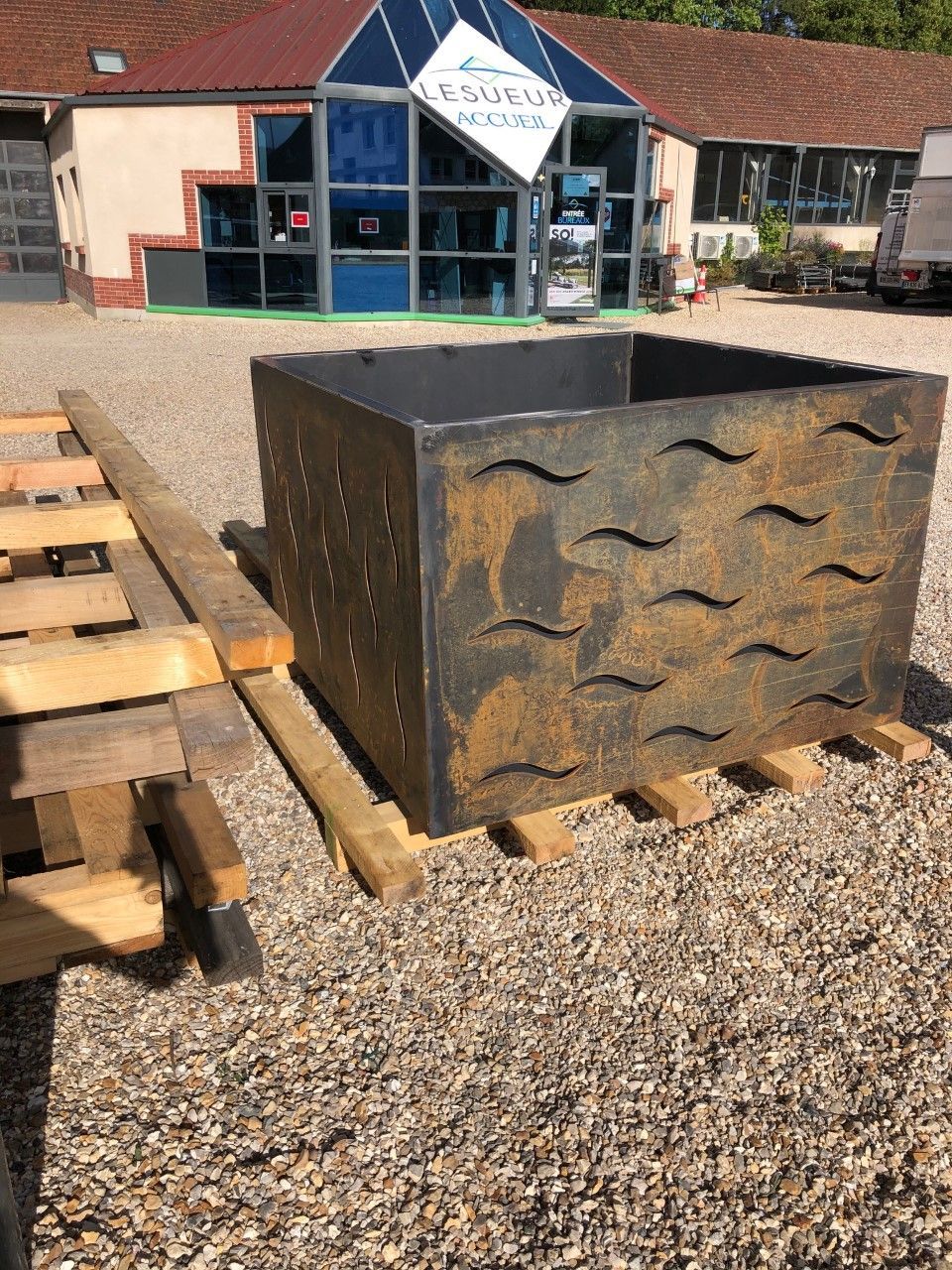 Grande sculpture cubique en métal rouillé posée sur du gravier à côté de palettes en bois empilées près d'un bâtiment