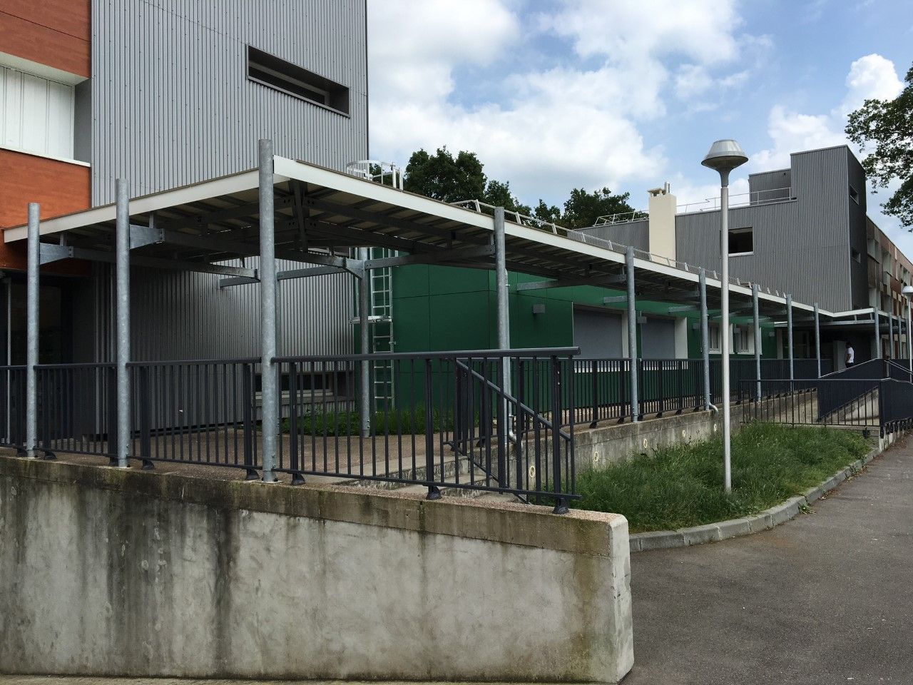 Passage couvert longeant un bâtiment gris, avec des rambardes métalliques et un mur en béton sous un ciel nuageux