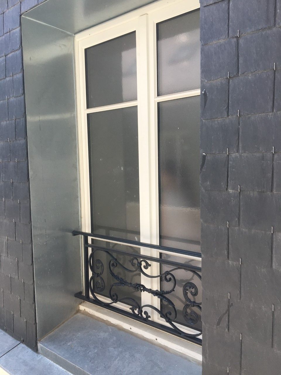Haute fenêtre à cadre blanc avec une rambarde en fer forgé noir sur une façade d'immeuble en pierre grise