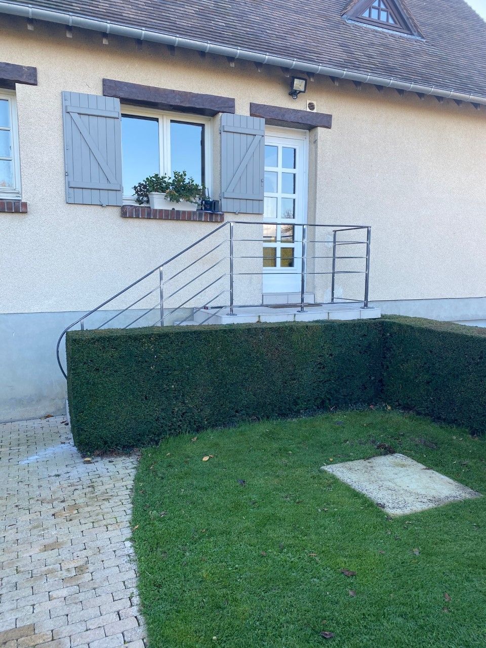 Entrée de la maison avec volets bleus, porte blanche, rampe d'accès en métal et haie taillée dans la cour avant