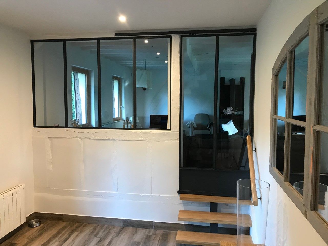 Pièce vide aux murs blancs, parquet, fenêtres à cadre noir et un petit escalier près d'une porte vitrée