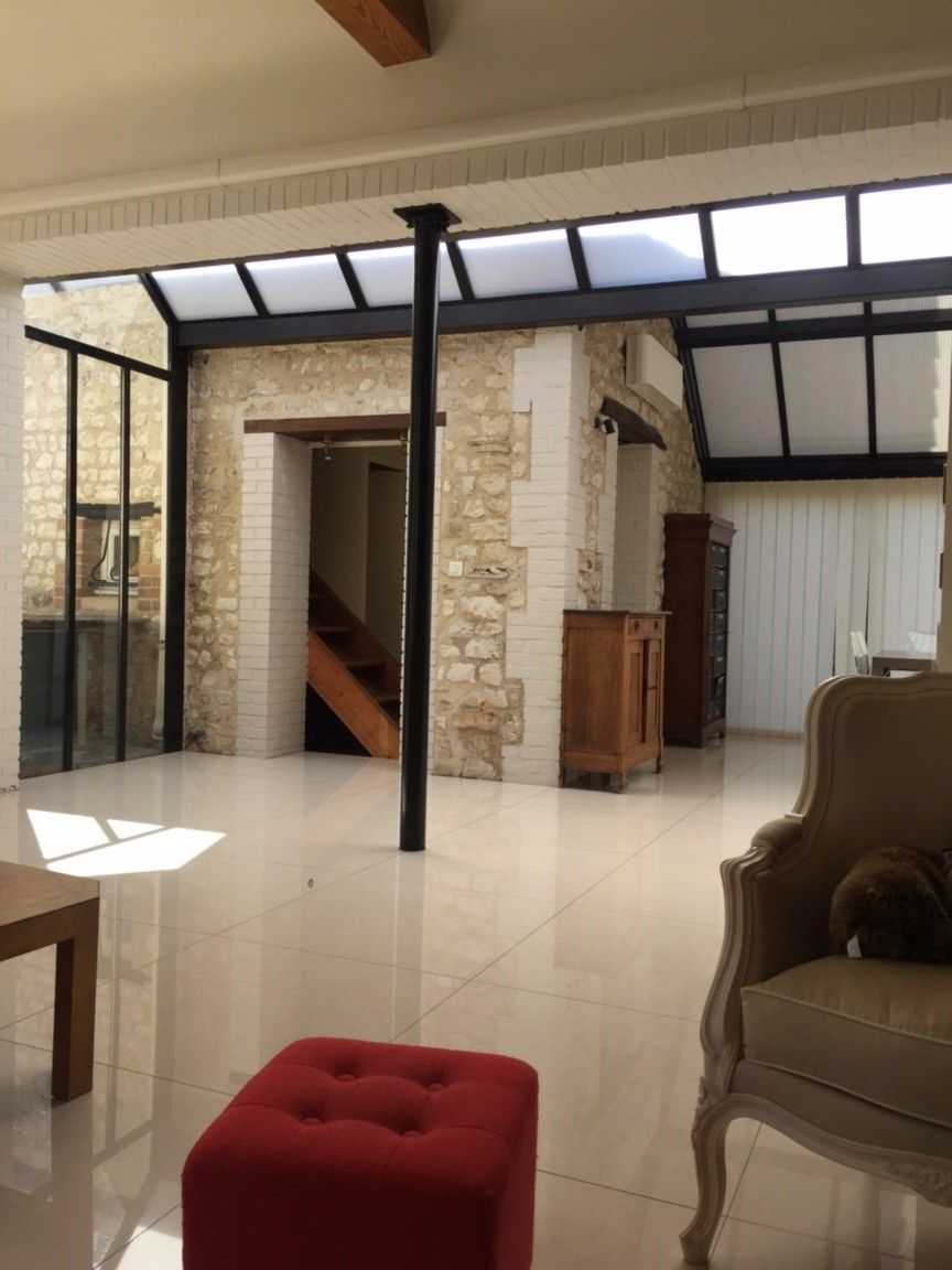 Patio intérieur lumineux avec murs en pierre, poteaux de soutien noirs, sol carrelé et rideaux blancs ; pouf et fauteuil rouges au premier plan