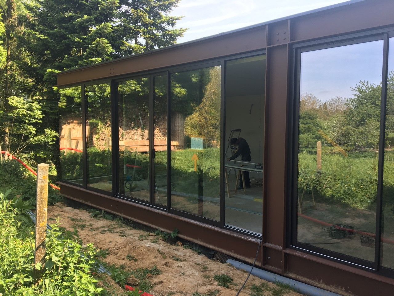 Véranda moderne en verre aux cadres sombres, nichée au milieu des arbres et de la pelouse d'un jardin.