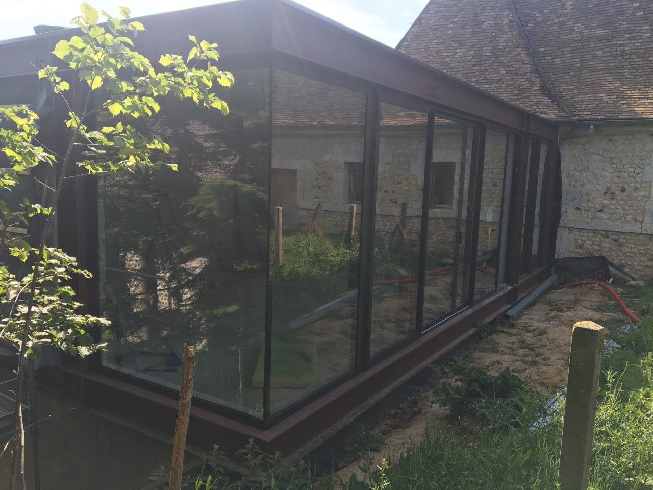 Véranda vitrée jouxtant une maison en pierre, avec un jeune arbre au premier plan.