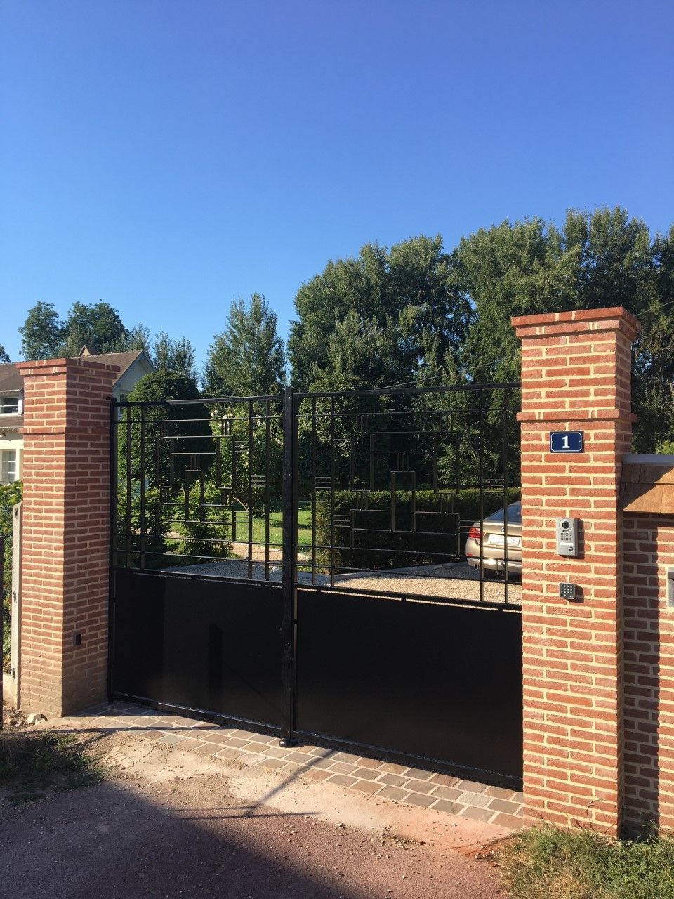 Entrée fermée par un portail en briques et en métal, avec des piliers en briques et des arbres à l'arrière, sous un ciel bleu clair.