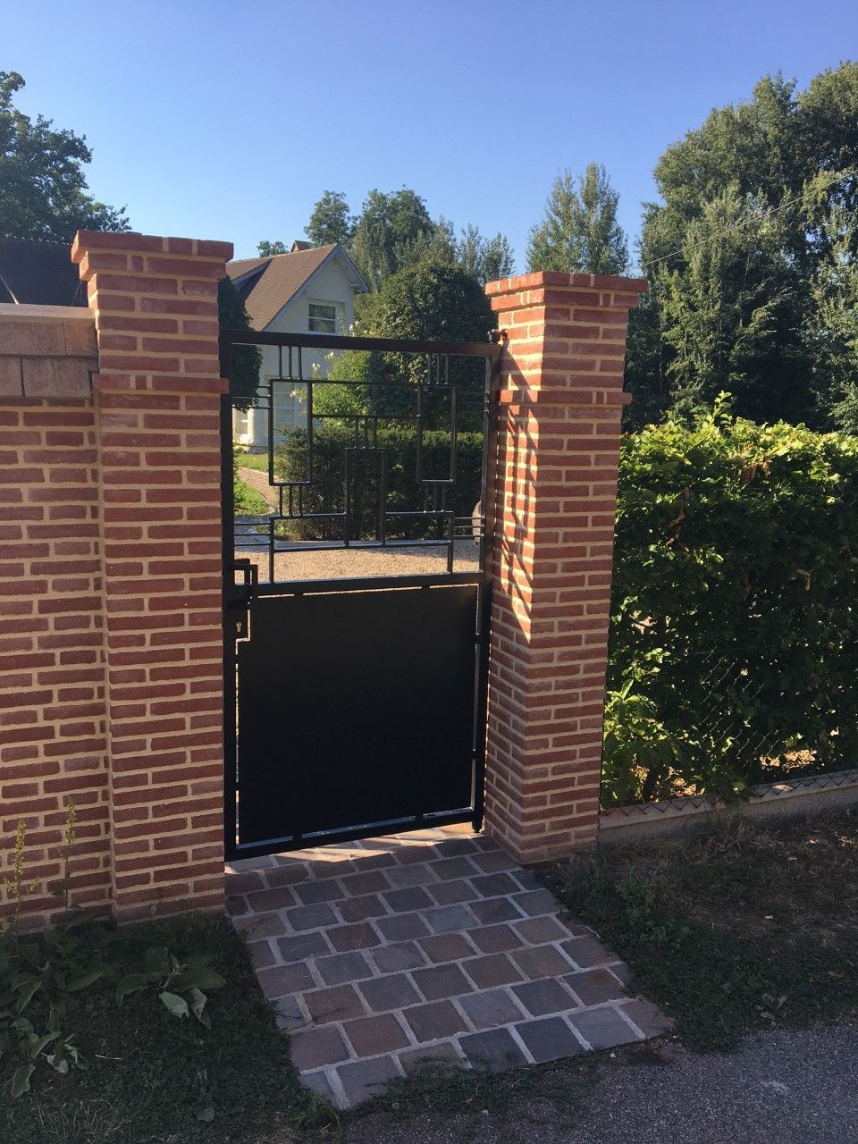 Portail de jardin en briques avec panneaux métalliques noirs et allée en briques permettant de le traverser.