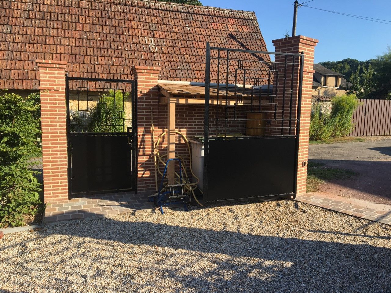 Entrée d'une maison en briques avec portail en métal noir et allée de gravier sous un ciel ensoleillé