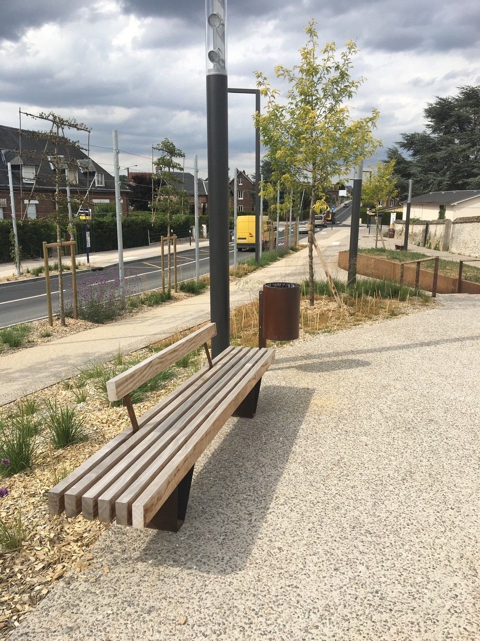 Long banc en bois au bord d'une rue tranquille et d'une piste cyclable, avec de jeunes arbres et un ciel nuageux.