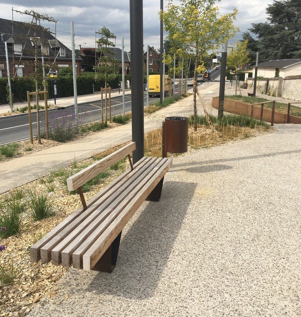 Banc en bois au bord d'un chemin de gravier dans un paysage urbain moderne, avec de jeunes arbres et une voie ferrée à proximité.